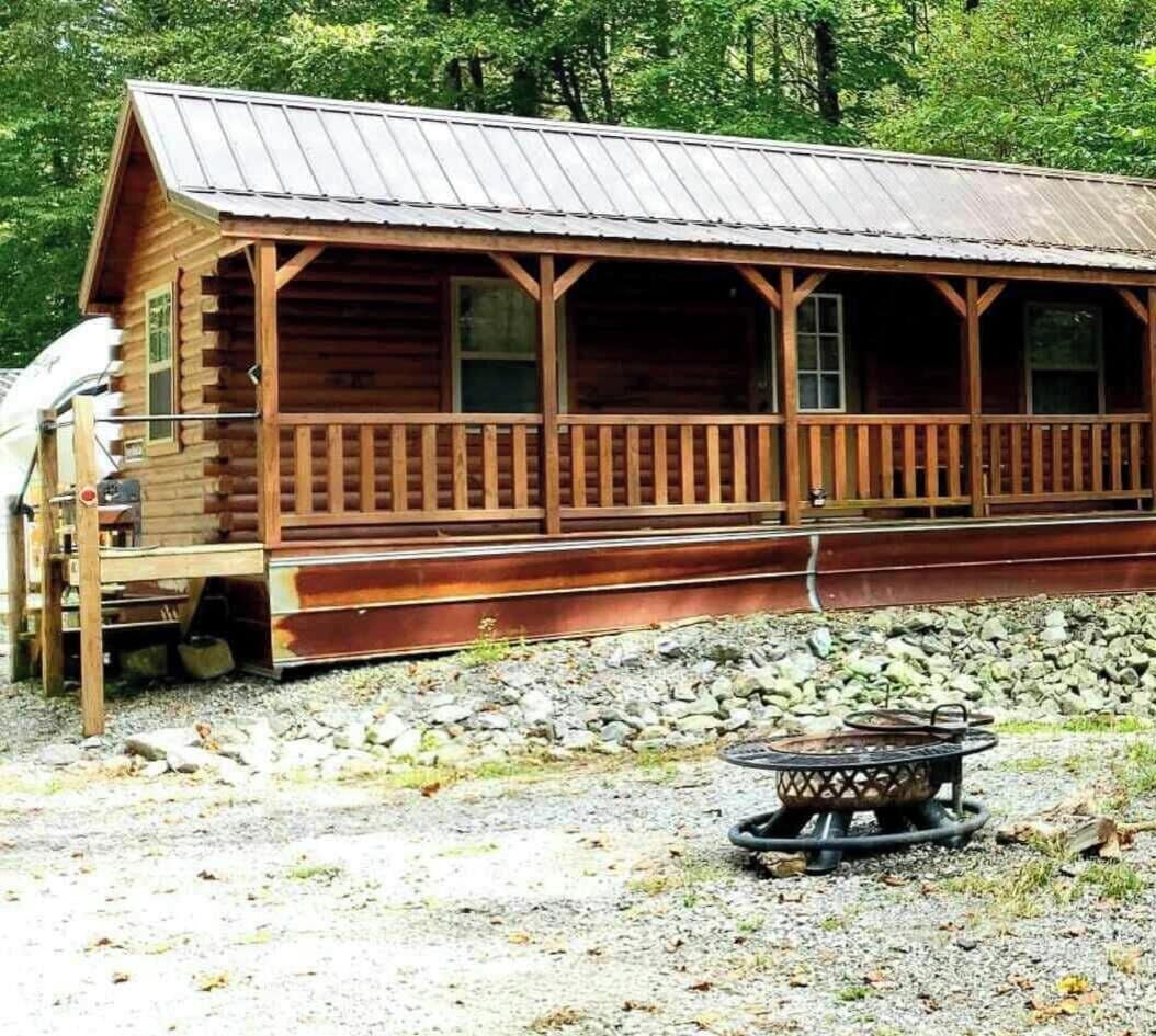 A rustic wooden cabin with a porch, a fire pit, and a gravel ground.