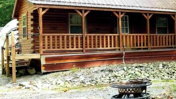 A rustic wooden cabin with a porch, a fire pit, and a gravel ground.