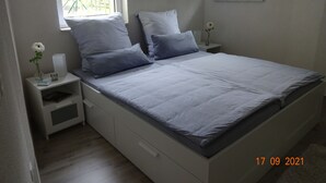Une chambre à coucher comprenant un lit, deux tables de chevet, une commode, une fenêtre et une petite table sur laquelle est posé un vase de fleurs.