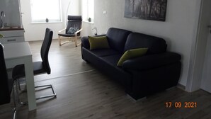 Un salon moderne avec un canapé noir, une table à manger et une chaise. Un tableau orne le mur, et une fenêtre offre une vue sur les arbres.