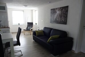 Een moderne woonkamer met een zwarte bank, een eettafel en een stoel. Er hangt een schilderij aan de muur en een raam met uitzicht op bomen.