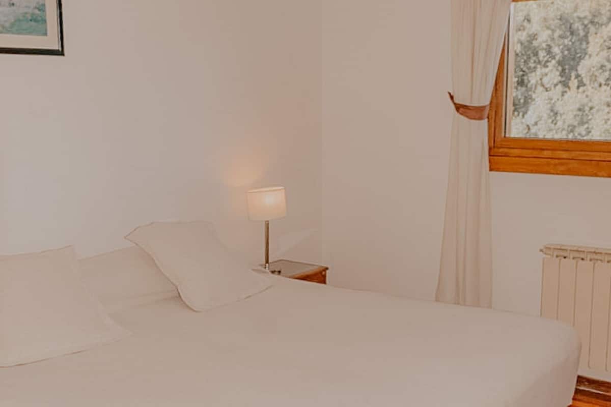 Un dormitorio con una cama grande, una mesita de noche con lámpara, una ventana con cortinas y un radiador.