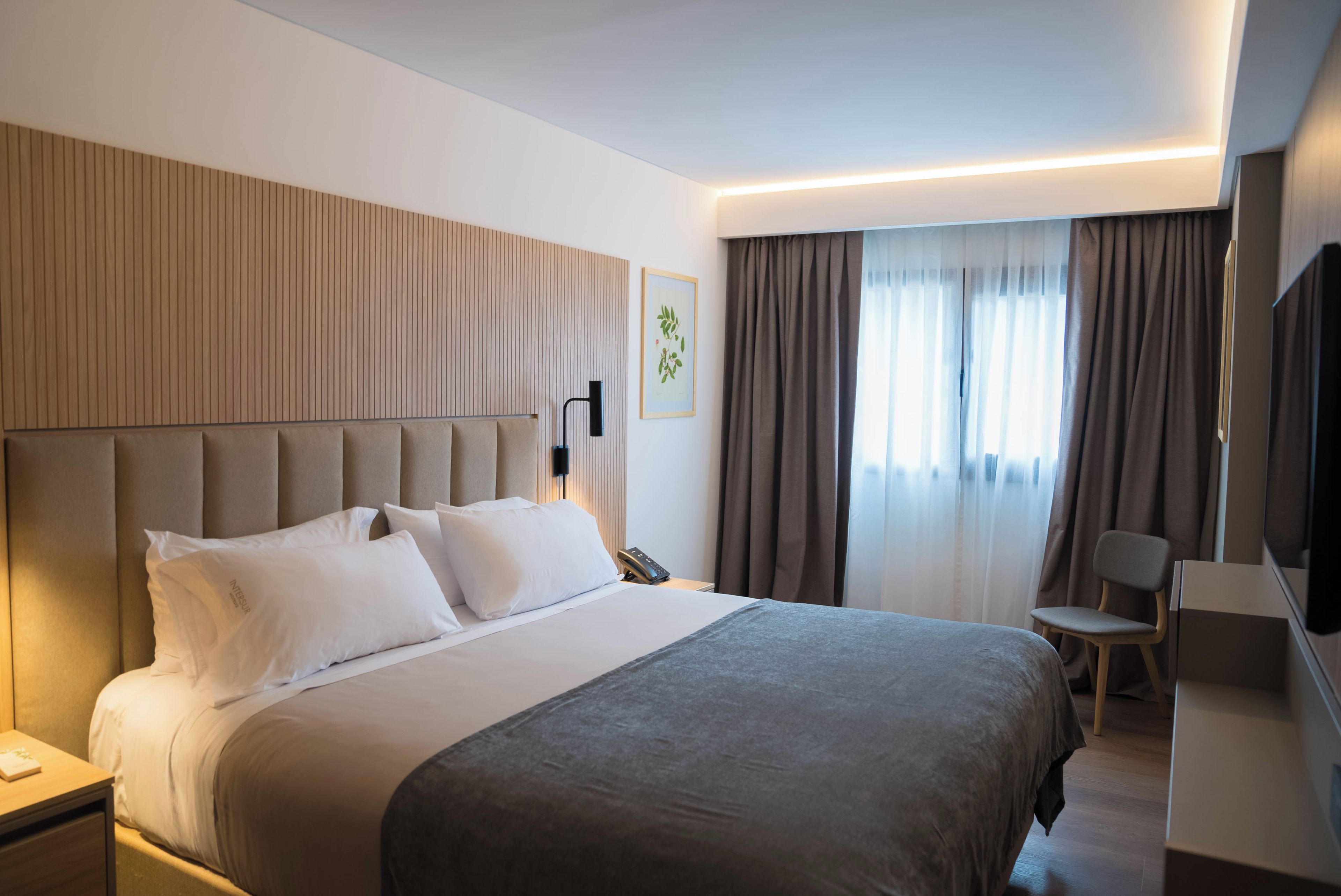 Habitación de hotel con una cama grande, una mesita de noche, un escritorio con silla, un televisor de pantalla plana y una ventana con cortinas.