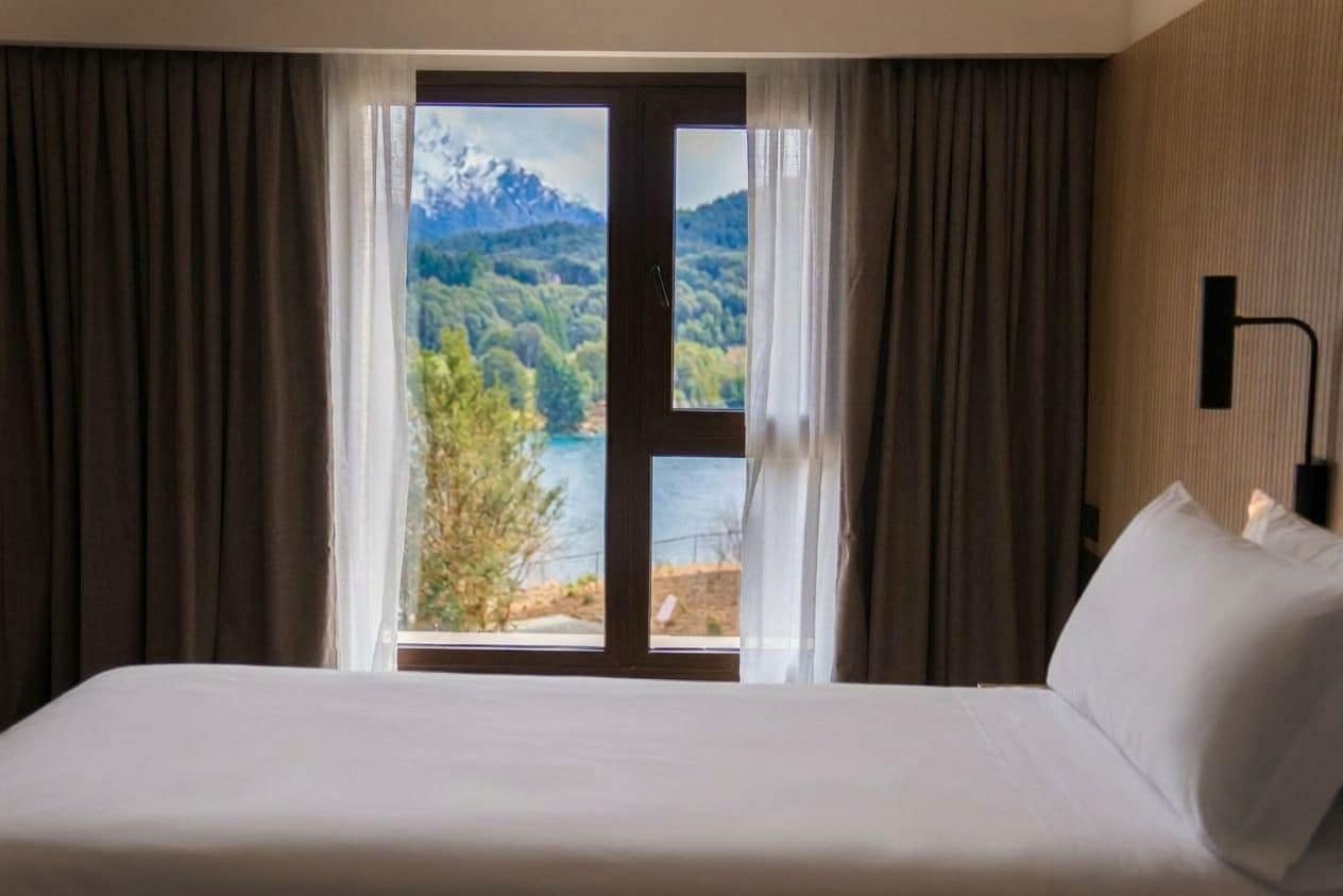 Een hotelkamer met een bed, uitzicht op een meer en bergen, en een deur met gordijnen.
