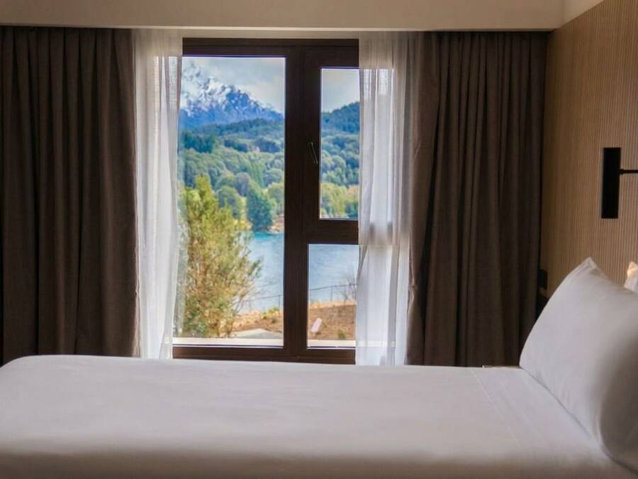 Een hotelkamer met een bed, uitzicht op een meer en bergen, en een deur met gordijnen.