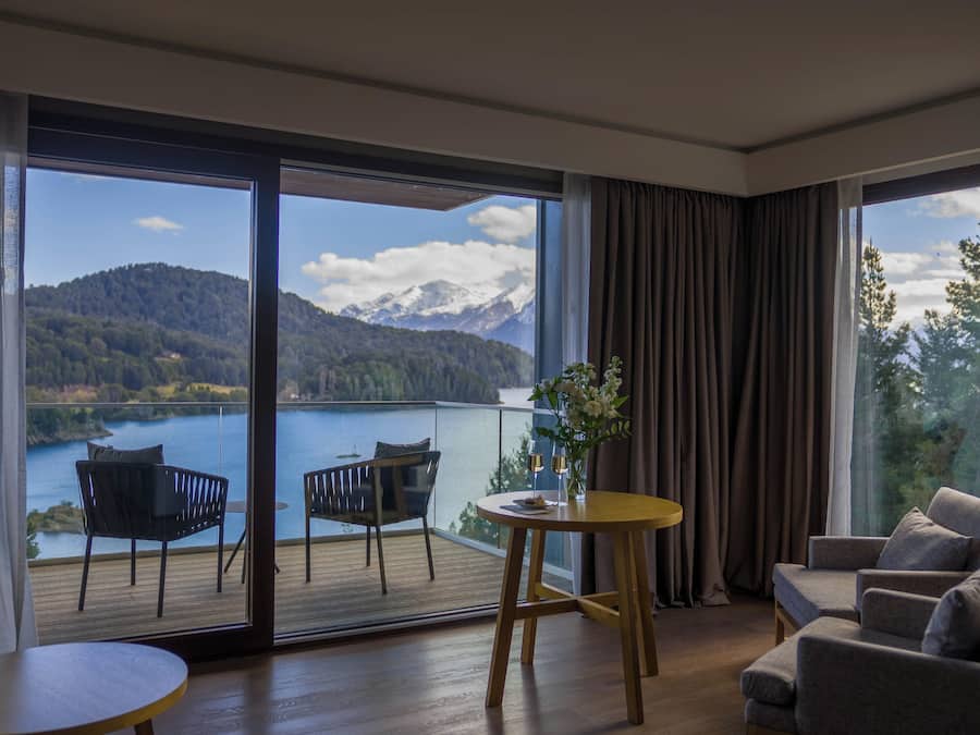 Habitación con un amplio ventanal corredizo que da a un balcón con vistas a un lago y montañas. En el interior, se encuentra una mesa redonda de madera con dos sillas y un sofá.