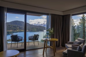 A room with a large sliding glass door leading to a balcony overlooking a lake and mountains. Inside, there is a round wooden table with two chairs and a sofa.