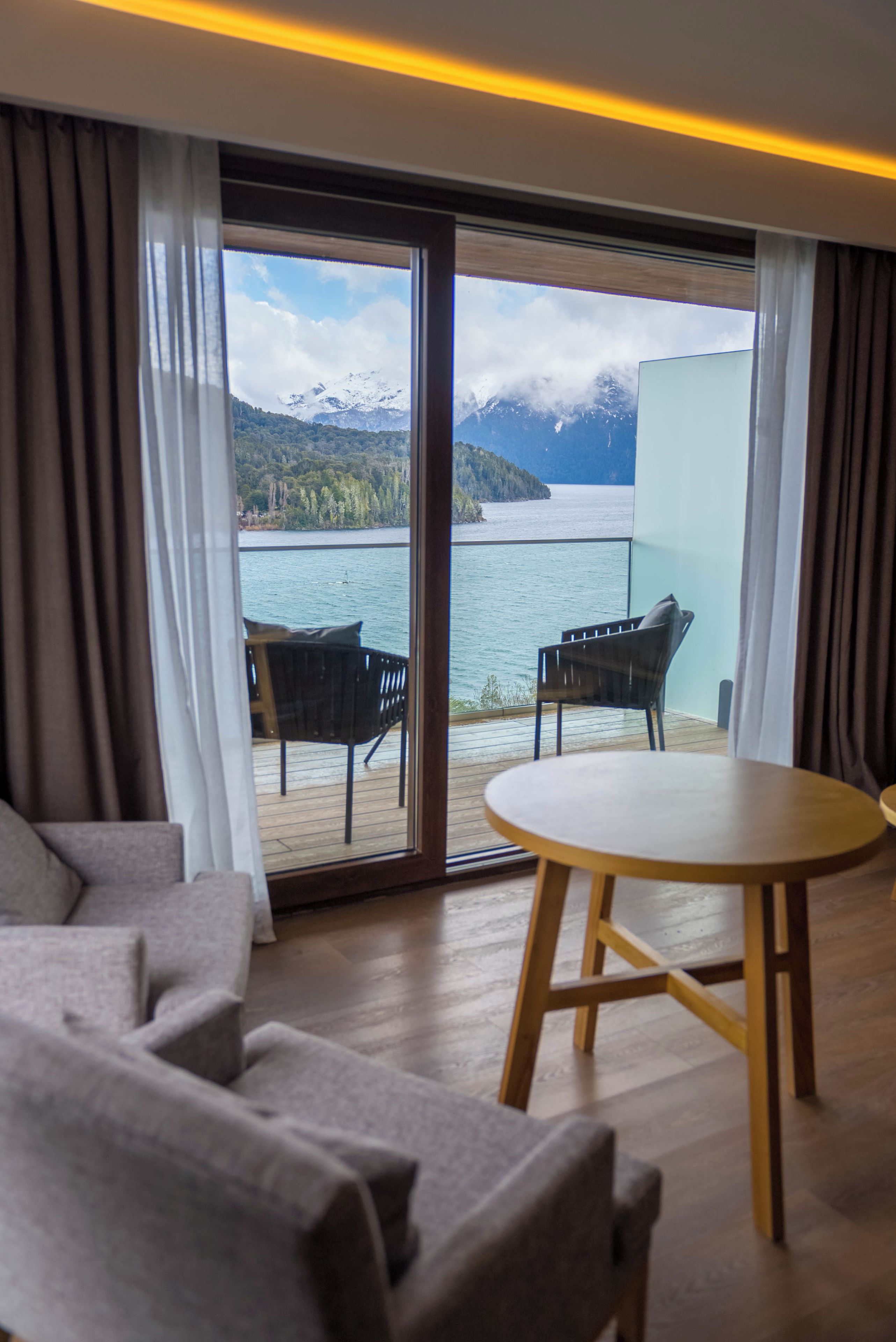 A room with a sofa, a table, and a view of a lake and mountains through a glass door.