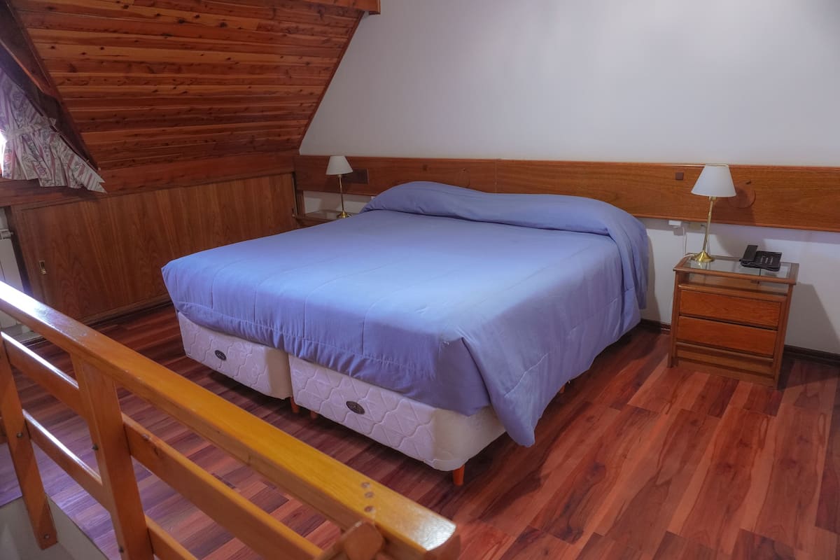 Un dormitorio con techo de madera, una cama con colcha azul, una mesita de noche con lámpara y un banco de madera.