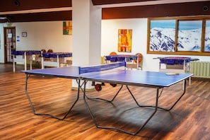 Una mesa de ping pong en una habitación con suelo de madera, un cuadro de un paisaje montañoso y una ventana con vistas a picos nevados.