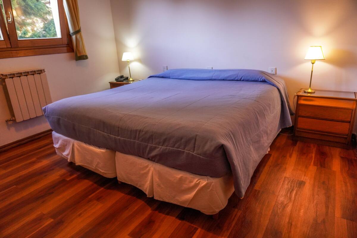 Un dormitorio con una cama grande, suelo de madera, dos mesitas de noche con lámparas y una ventana con cortinas.