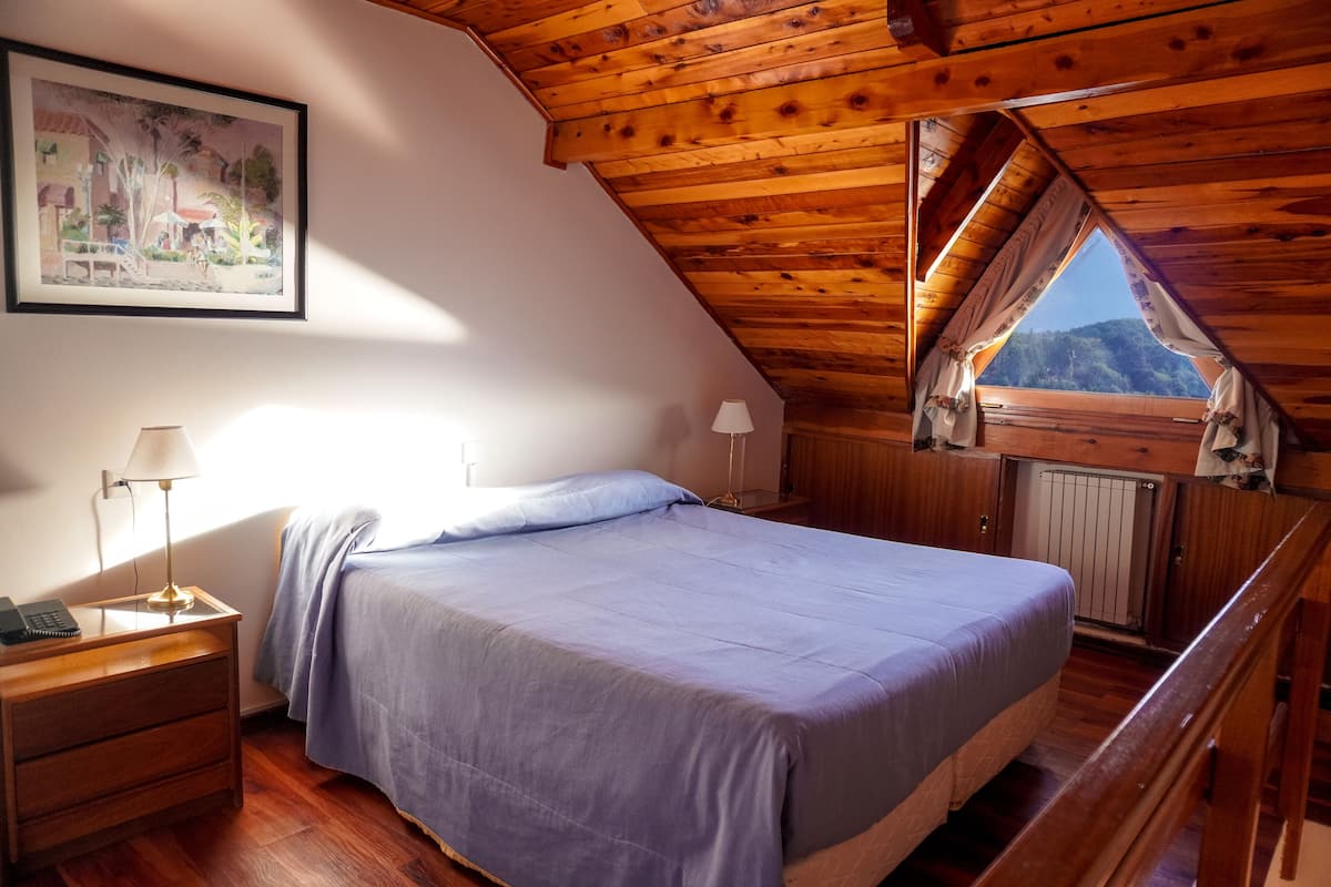 Un dormitorio con techo de madera, una cama con colcha azul, una mesita de noche con lámpara y una ventana con cortinas.