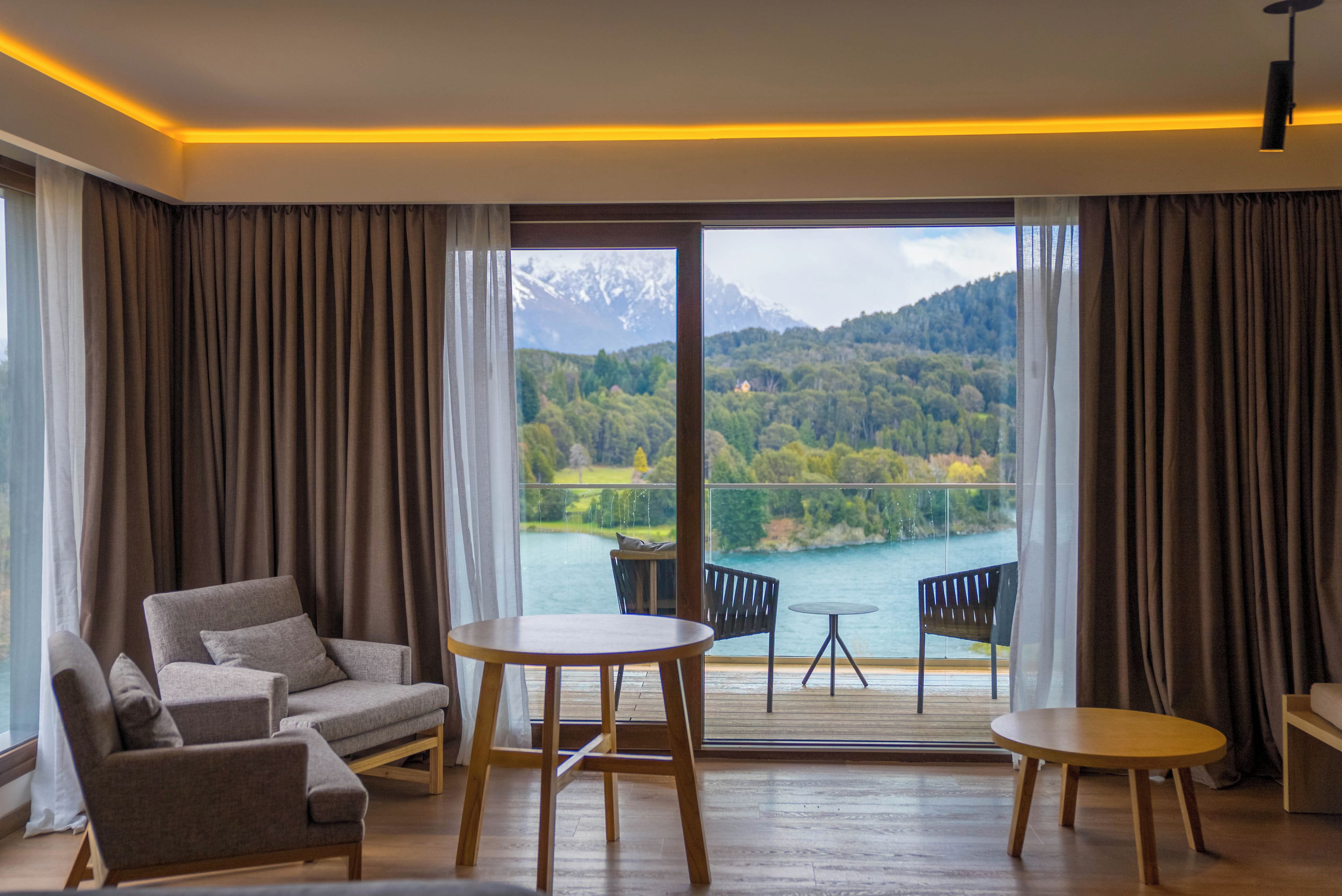 Una habitación de hotel moderna con sofá, dos mesas y vistas a un lago y montañas.