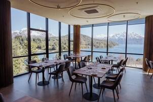Comedor con mesas dispuestas para recibir a los huéspedes, amplias ventanas con vistas a montañas y un lago.