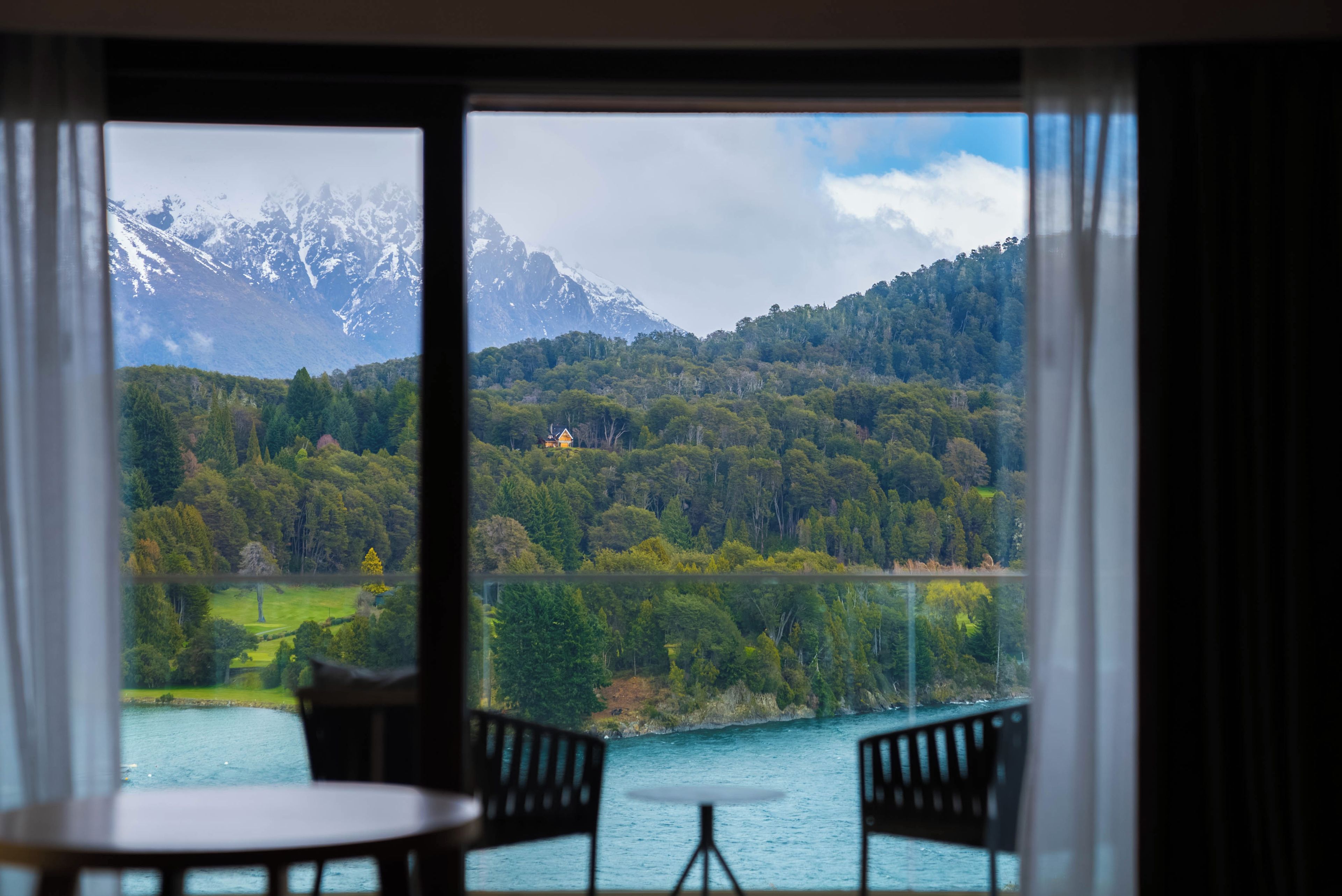 Une pièce avec vue sur un lac, des montagnes et une forêt. On y aperçoit deux chaises et une table au premier plan.