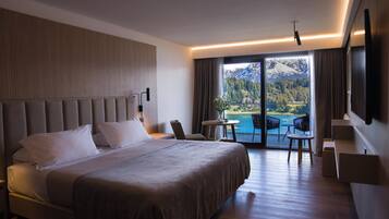 Une chambre d’hôtel moderne dotée d’un grand lit, d’un coin salon avec une table et des chaises, et d’un balcon offrant une vue panoramique sur les montagnes.