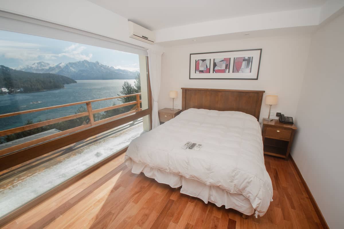 Un dormitorio con un ventanal grande que ofrece vistas a un lago y montañas. Hay una cama con edredón blanco, dos mesitas de noche y un cuadro enmarcado en la pared.