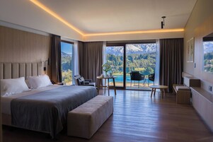 A modern hotel room with a large bed, a seating area with a table, and a balcony with a mountain view.
