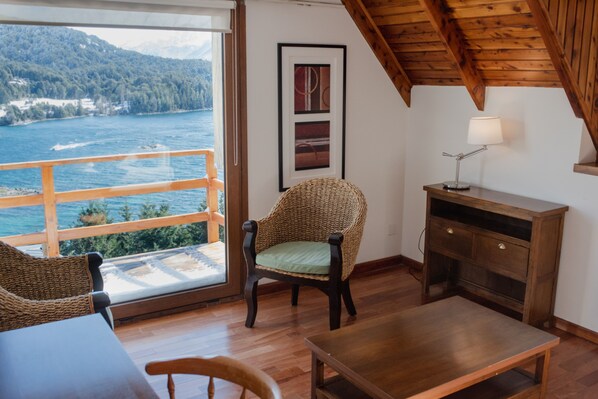 Quarto com uma mesa de madeira, uma cadeira de vime, um armário de madeira e vista para um lago e montanhas.