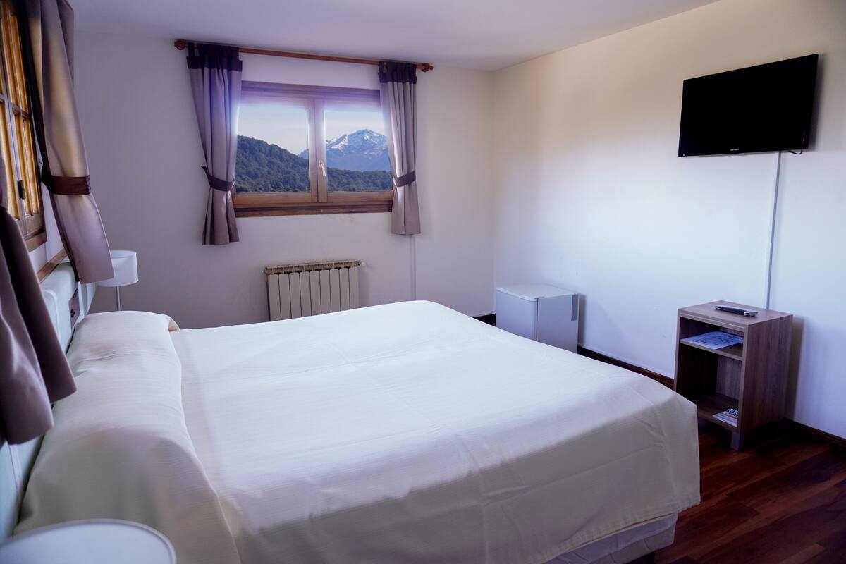 Una cama bien tendida con vistas a las montañas a través de una ventana, un televisor de pantalla plana en la pared y una pequeña mesita de noche con un control remoto.