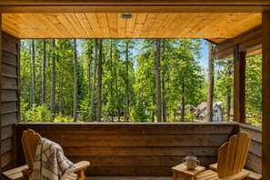 Puinen mökki, jossa on kuisti, kaksi tuolia, pöytä ja näkymä metsään.