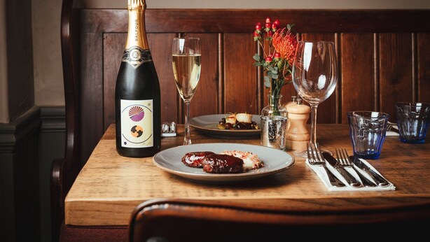 A wooden dining table with a bottle of champagne, a glass of white wine, a plate with food, and a vase with flowers. - The Falcon Painswick (Stroud)