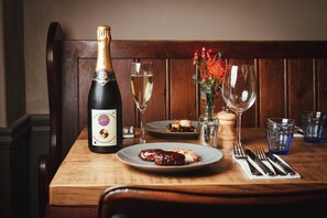 A wooden dining table with a bottle of champagne, a glass of white wine, a plate with food, and a vase with flowers. - The Falcon Painswick (Stroud)