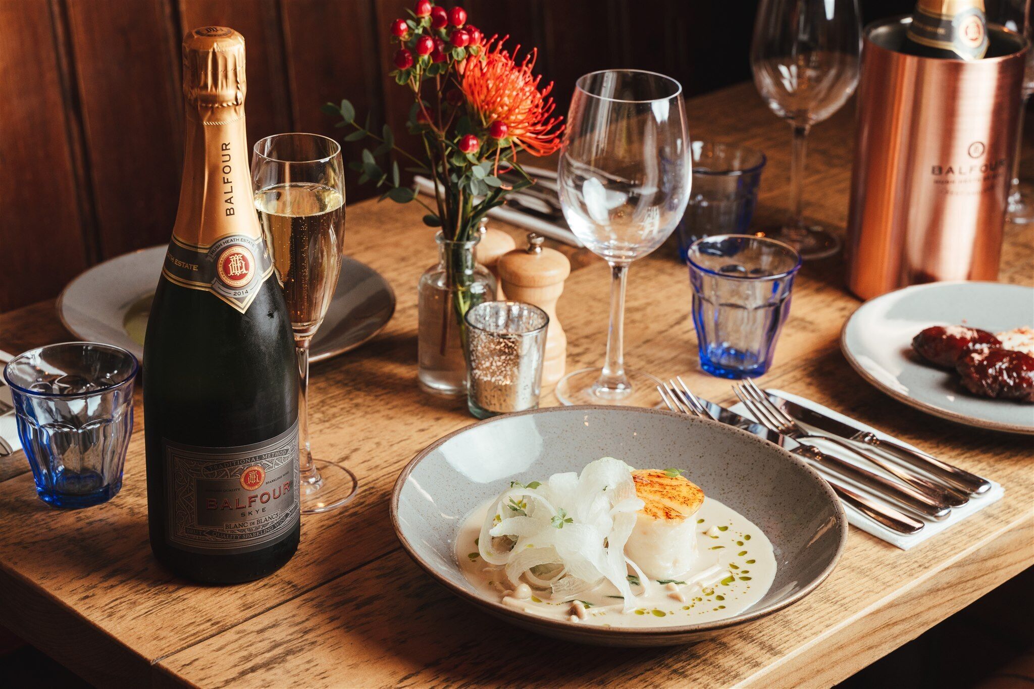 A table set with a bottle of Balfour champagne, a glass of water, a glass of wine, a copper decanter, a vase with red berries, a glass of water, a plate with a white fish dish, and cutlery.