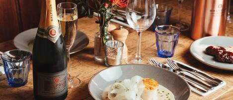 Una mesa dispuesta con una botella de champagne Balfour, un vaso de agua, una copa de vino, un decantador de cobre, un florero con bayas rojas, un vaso de agua, un plato con un guiso de pescado blanco y cubiertos.