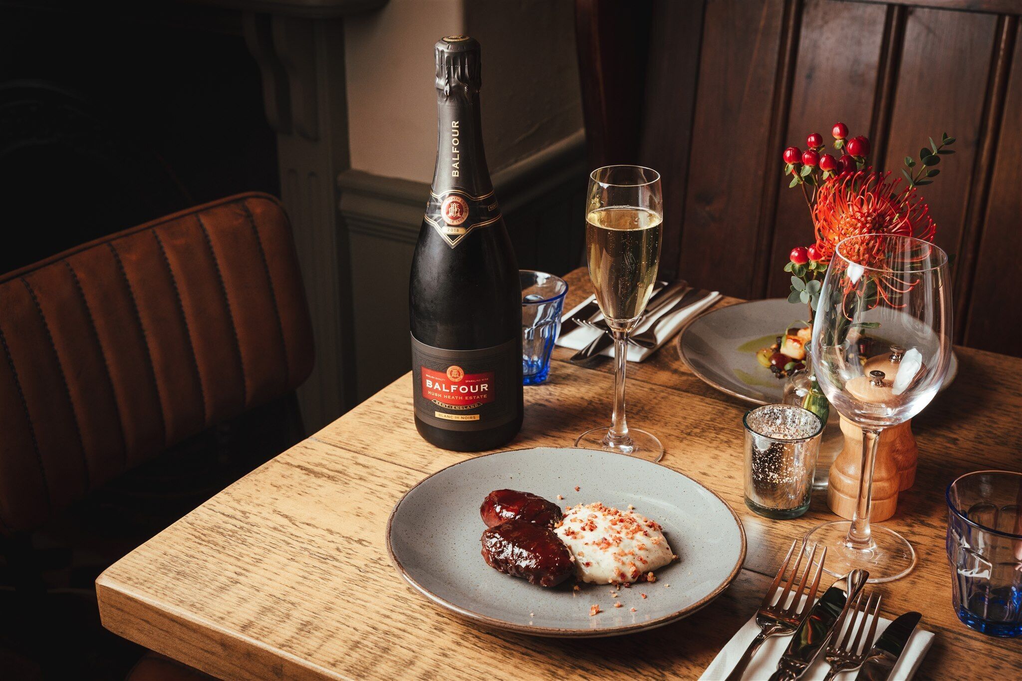 Une table en bois sur laquelle se trouvent une bouteille de champagne Balfour, un verre de champagne, une assiette avec de la nourriture et un verre agrémenté d’une composition décorative.