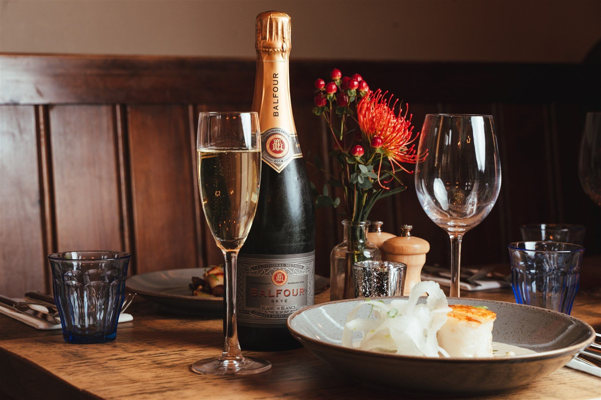 A bottle of Balfour champagne, two glasses, a bowl of food, and a decorative flower arrangement on a wooden table.