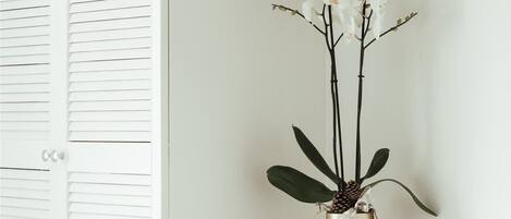 A white room with a white closet, a clear desk with a vase of white flowers, and a wall-mounted lamp.