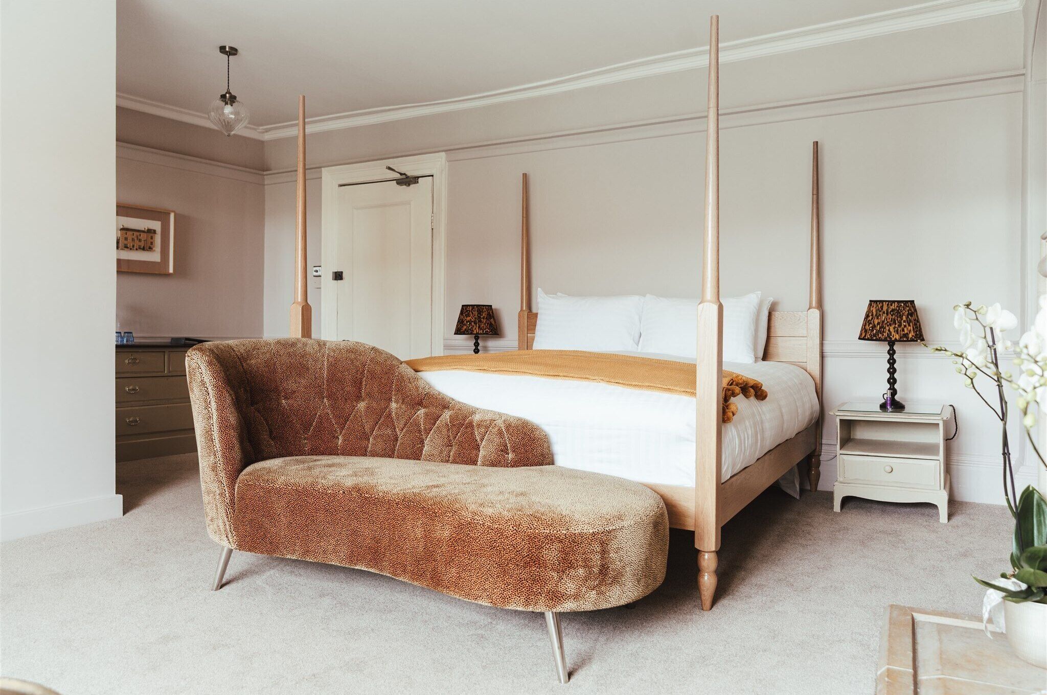 Une chambre à coucher avec un lit à baldaquin, un fauteuil marron recouvert de tissu, une table de chevet avec une lampe et une petite commode.