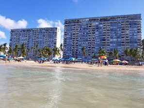 Un hotel frente a la playa con varios pisos, palmeras y sombrillas.