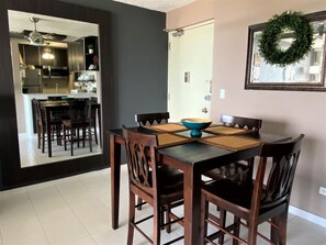 Un comedor con una mesa preparada para cuatro personas, rodeada de sillas de madera. Se ve una cocina al fondo, separada por un panel de vidrio.