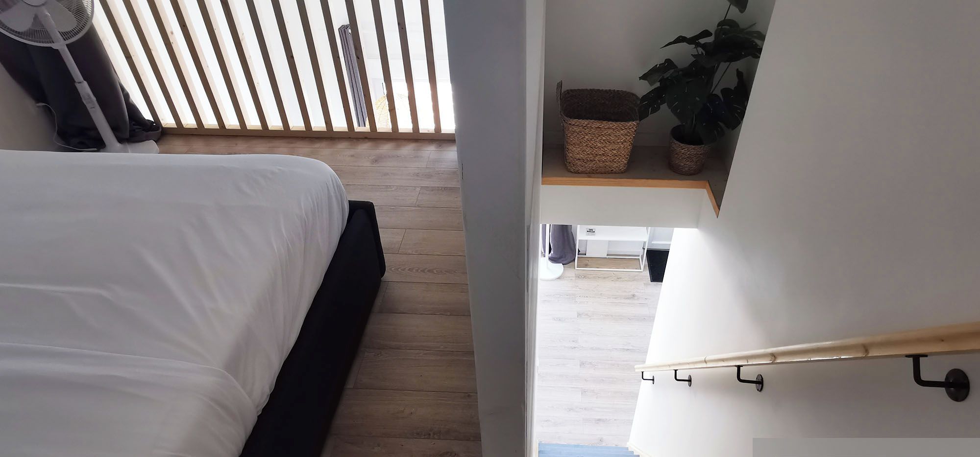 Een slaapkamer met een bed, houten vloeren, een ventilator en een trap naar een woonruimte.