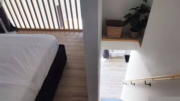 Een slaapkamer met een bed, houten vloeren, een ventilator en een trap naar een woonruimte.