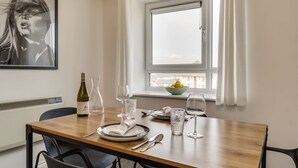 Ein Essbereich mit einem Holztisch, gedeckt für zwei Personen, einer Weinflasche, Weingläsern und einer Obstschale am Fensterbrett.