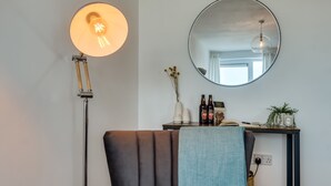Ein Zimmer mit einem gepolsterten Sessel, einem runden Spiegel, einer Stehlampe und einem Tisch mit einer Pflanze und zwei Bierflaschen.