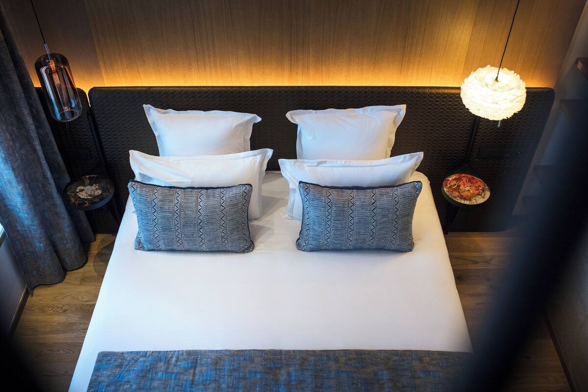 Una cama impecable con almohadas blancas y estampadas, una manta azul y un cabecero texturizado.