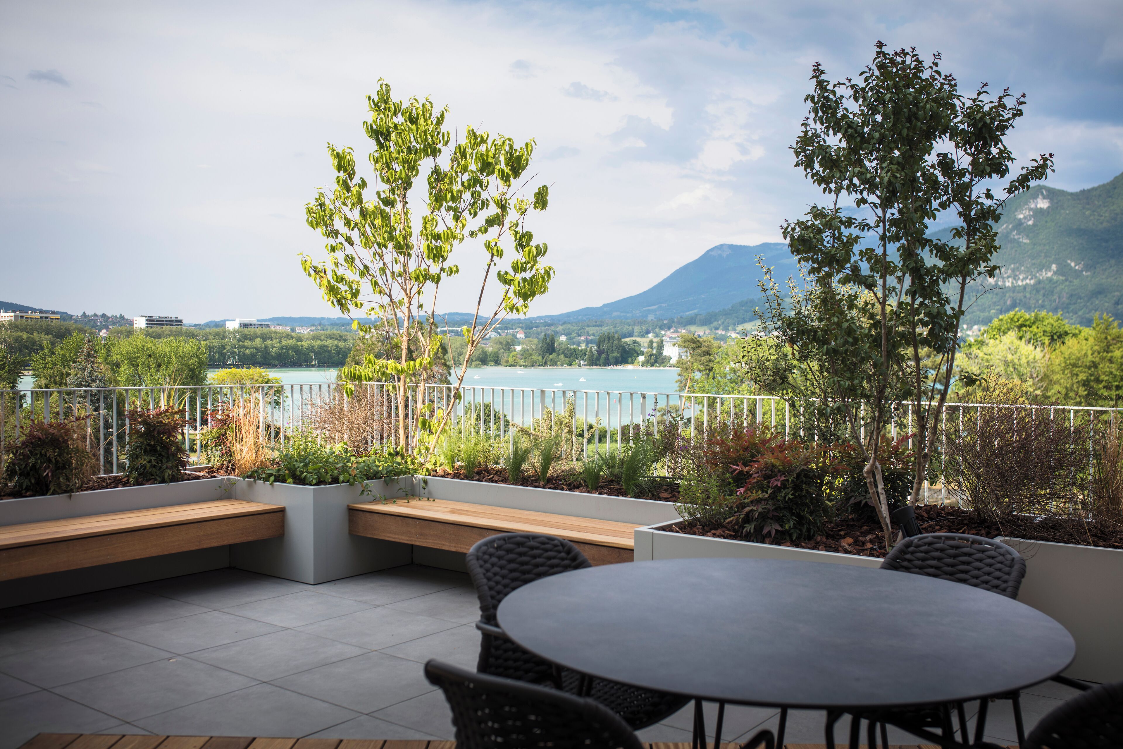 An outdoor seating area with a table and chairs, surrounded by plants and a view of a lake and mountains.