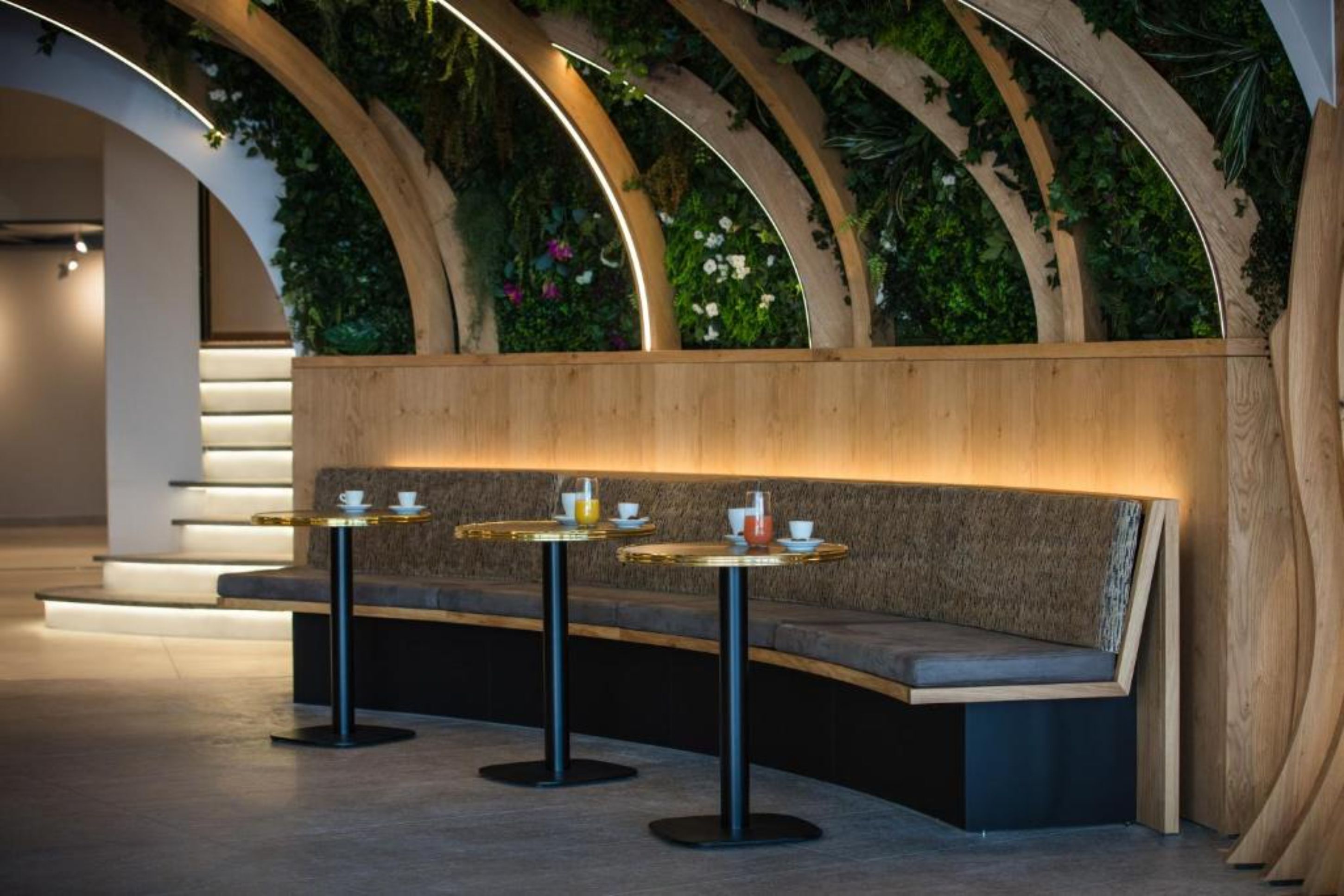Un área de descanso moderna con un banco curvo y mesas redondas, rodeada de arcos de madera y una pared verde con plantas.