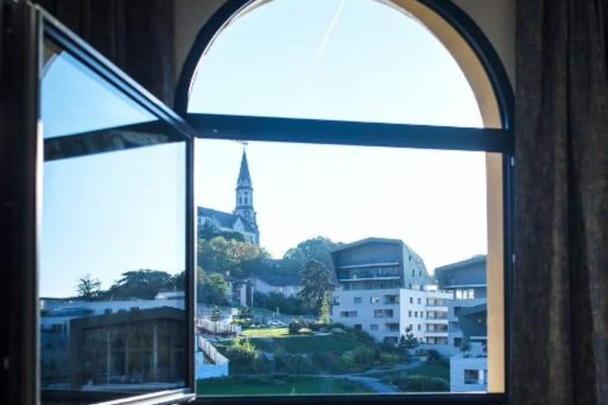 Una habitación con un amplio ventanal de arco, una cama con ropa oscura y vistas a una iglesia y edificios.