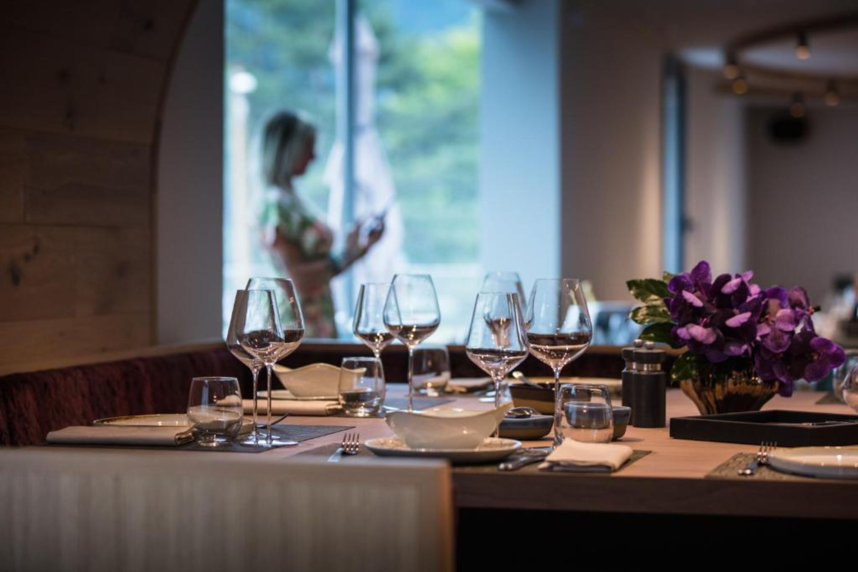 A dining table set with wine glasses, plates, and cutlery, with a vase of purple flowers and a view of a person outside.