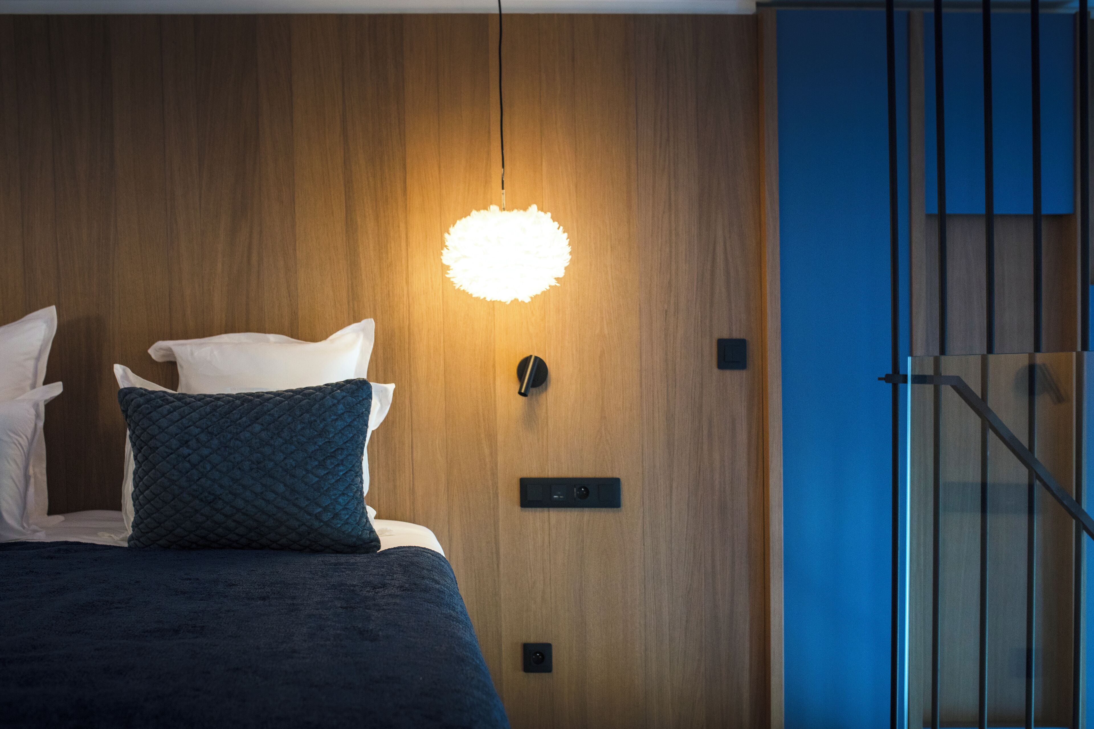 Un dormitorio moderno con pared de madera, cama con almohadas blancas y azul oscuro, una lámpara de techo circular original y una puerta azul.