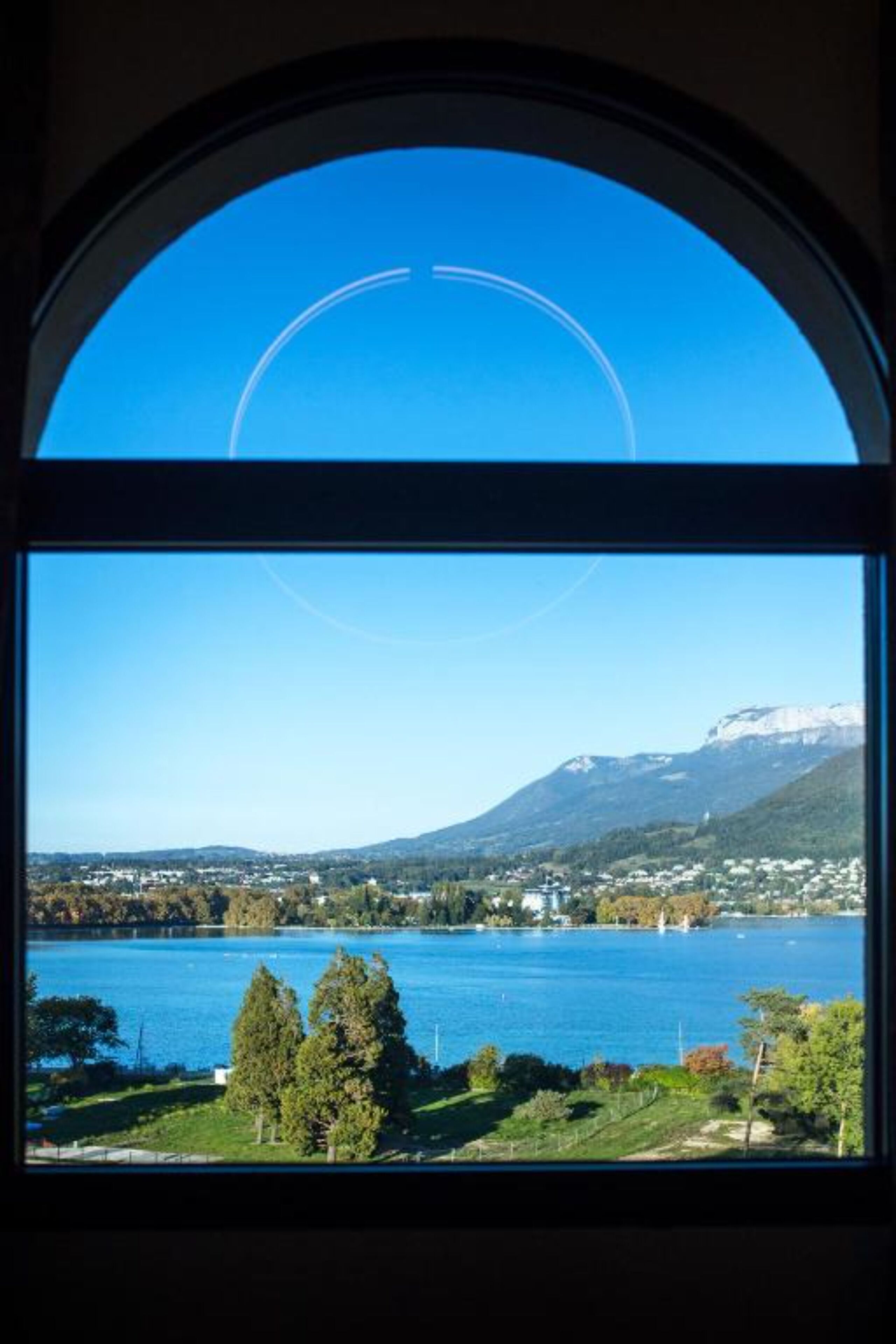Une vue à travers une fenêtre, avec un ciel bleu dégagé et un lac, des montagnes se dessinant à l’horizon.