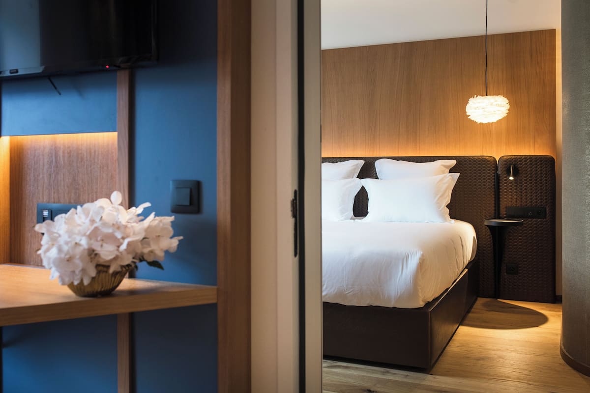 Una habitación de hotel moderna con una cama, una mesita de noche con un jarrón de flores, un televisor montado en la pared y una lámpara colgante original.