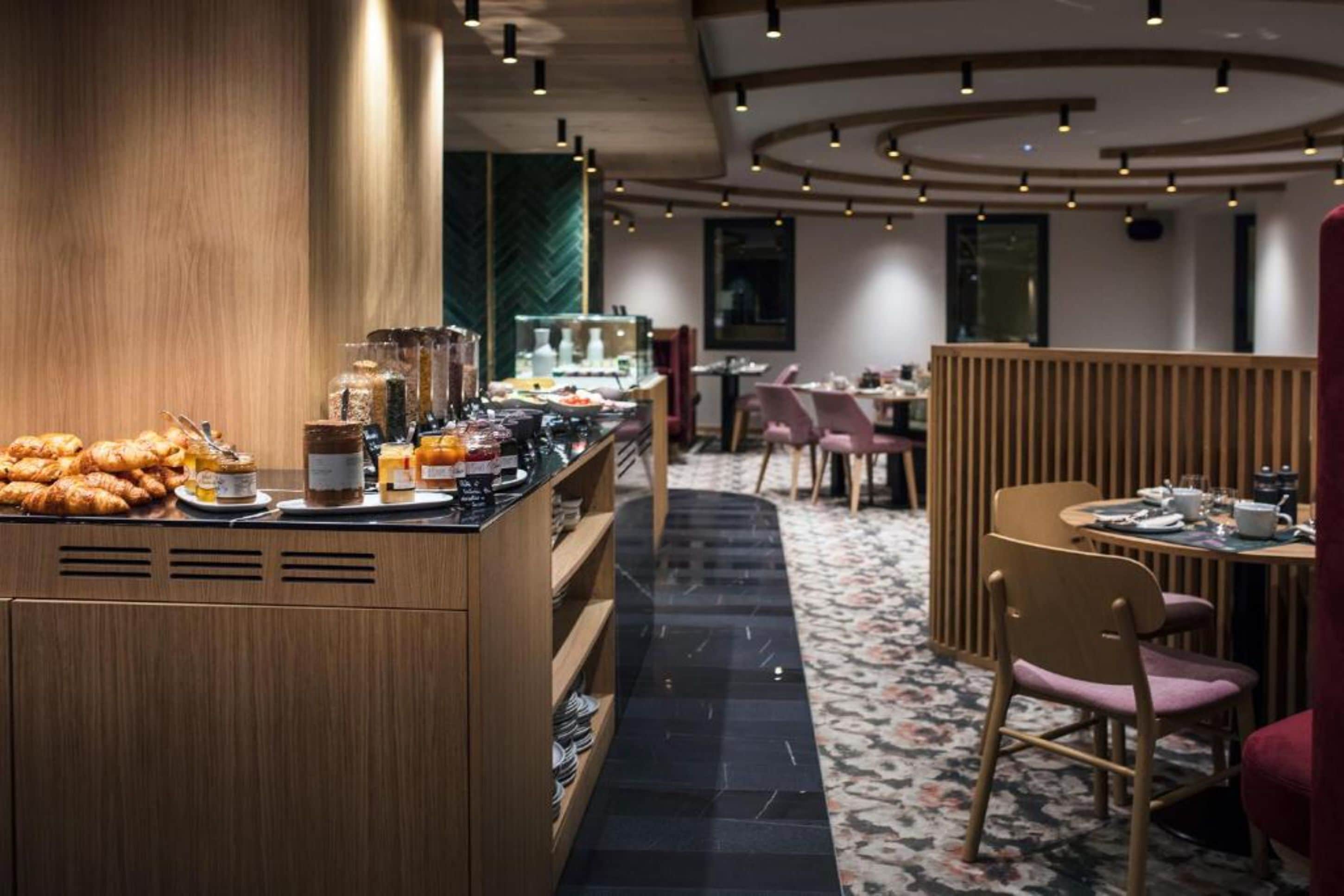 A hotel breakfast area with a wooden counter displaying pastries and beverages, a seating area with tables and chairs, and a patterned carpet.