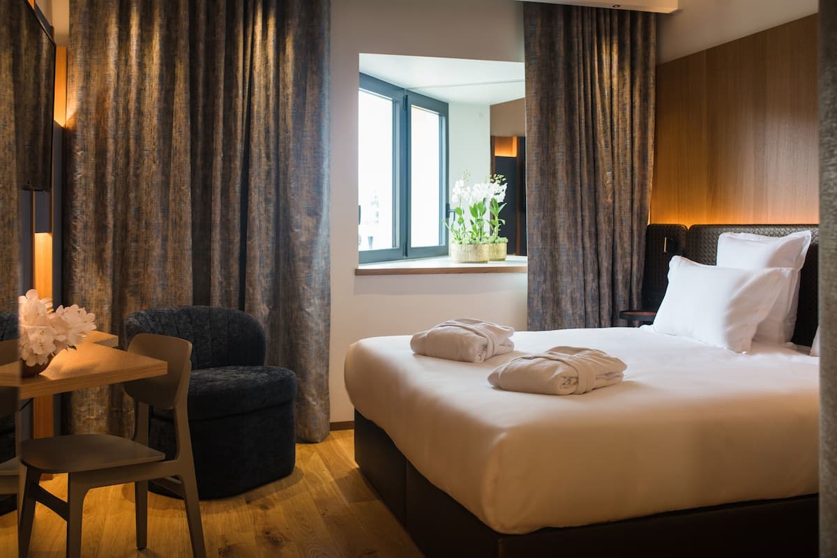 Una habitación de hotel con cama, escritorio con silla, ventana con cortinas y mesita de noche con lámpara.