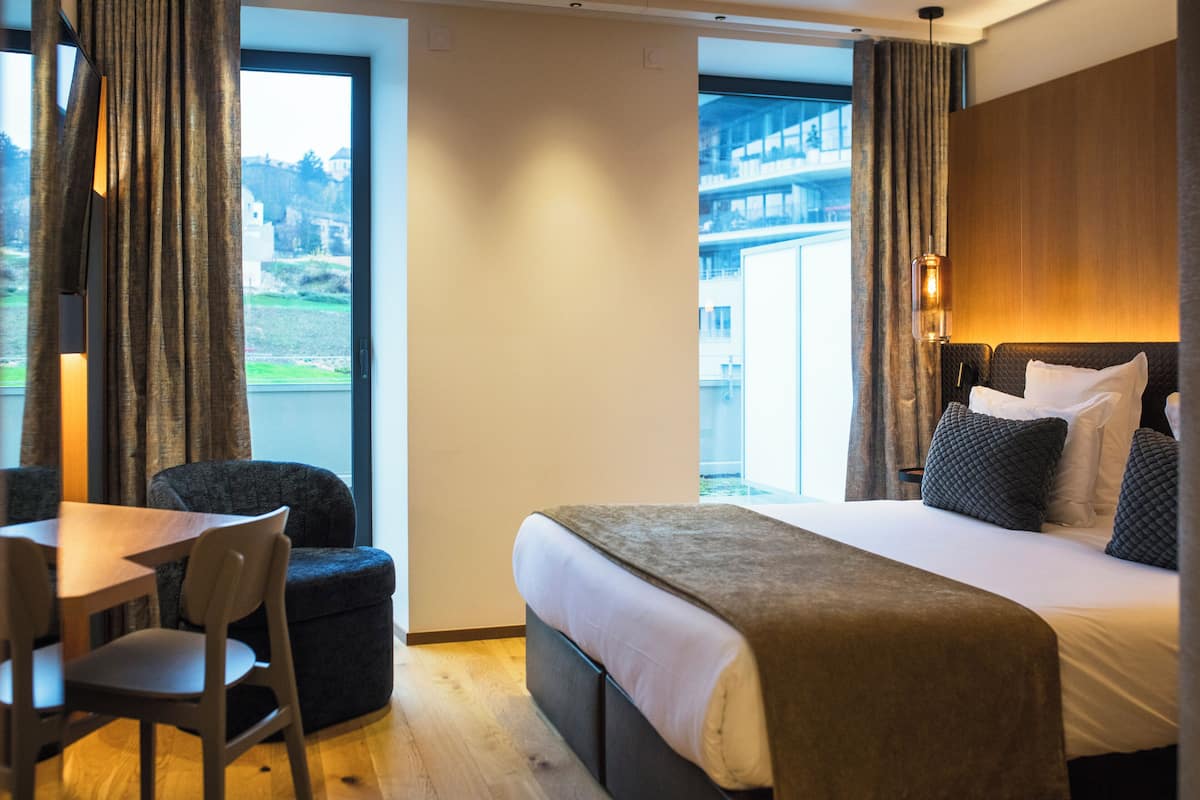 Habitación de hotel con cama, escritorio y sillas. Tiene una ventana con cortinas y vistas a edificios.